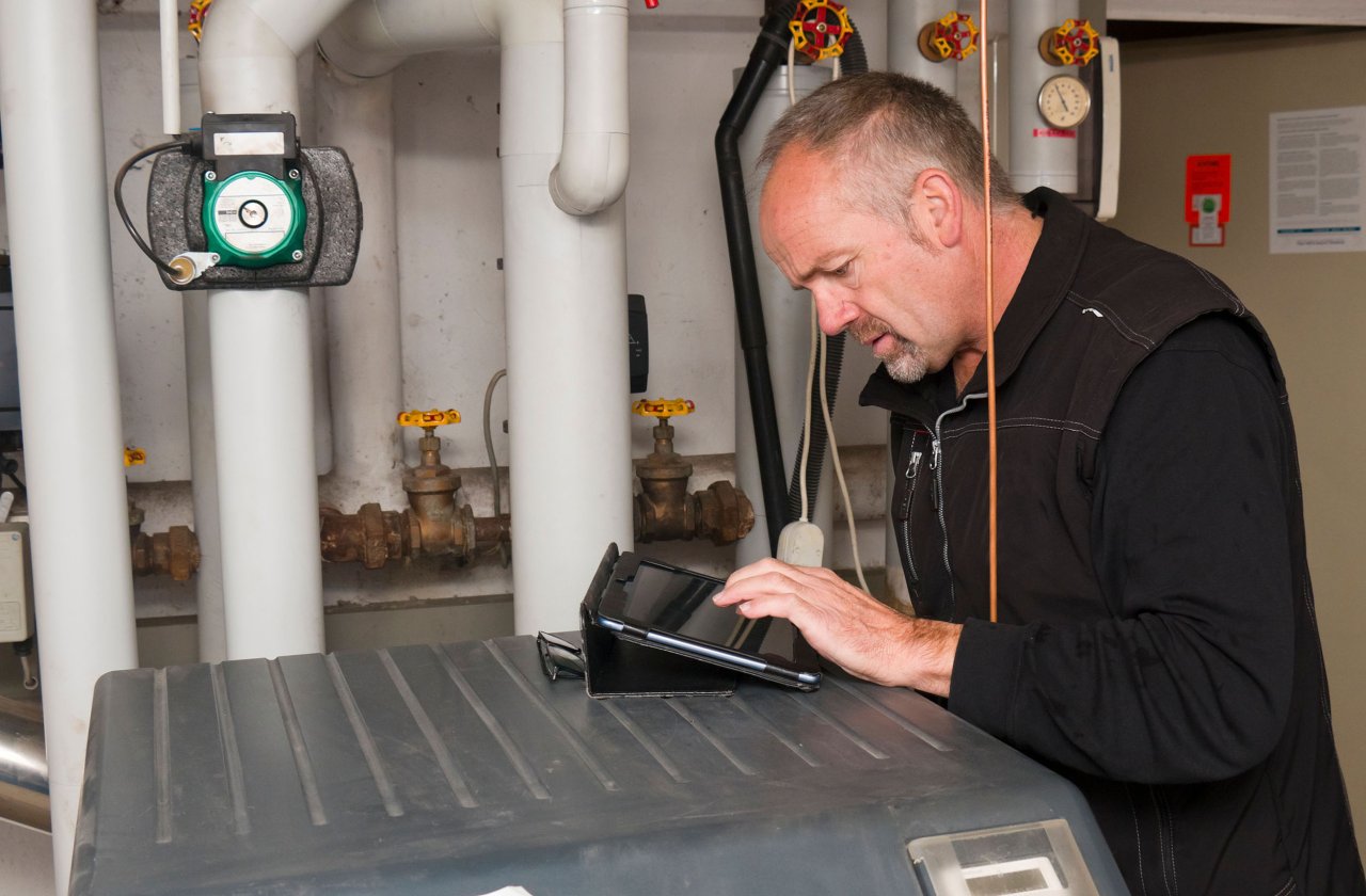 Techniker überprüft Heizungsanlage und bedient Tablet in einem Heizungsraum.