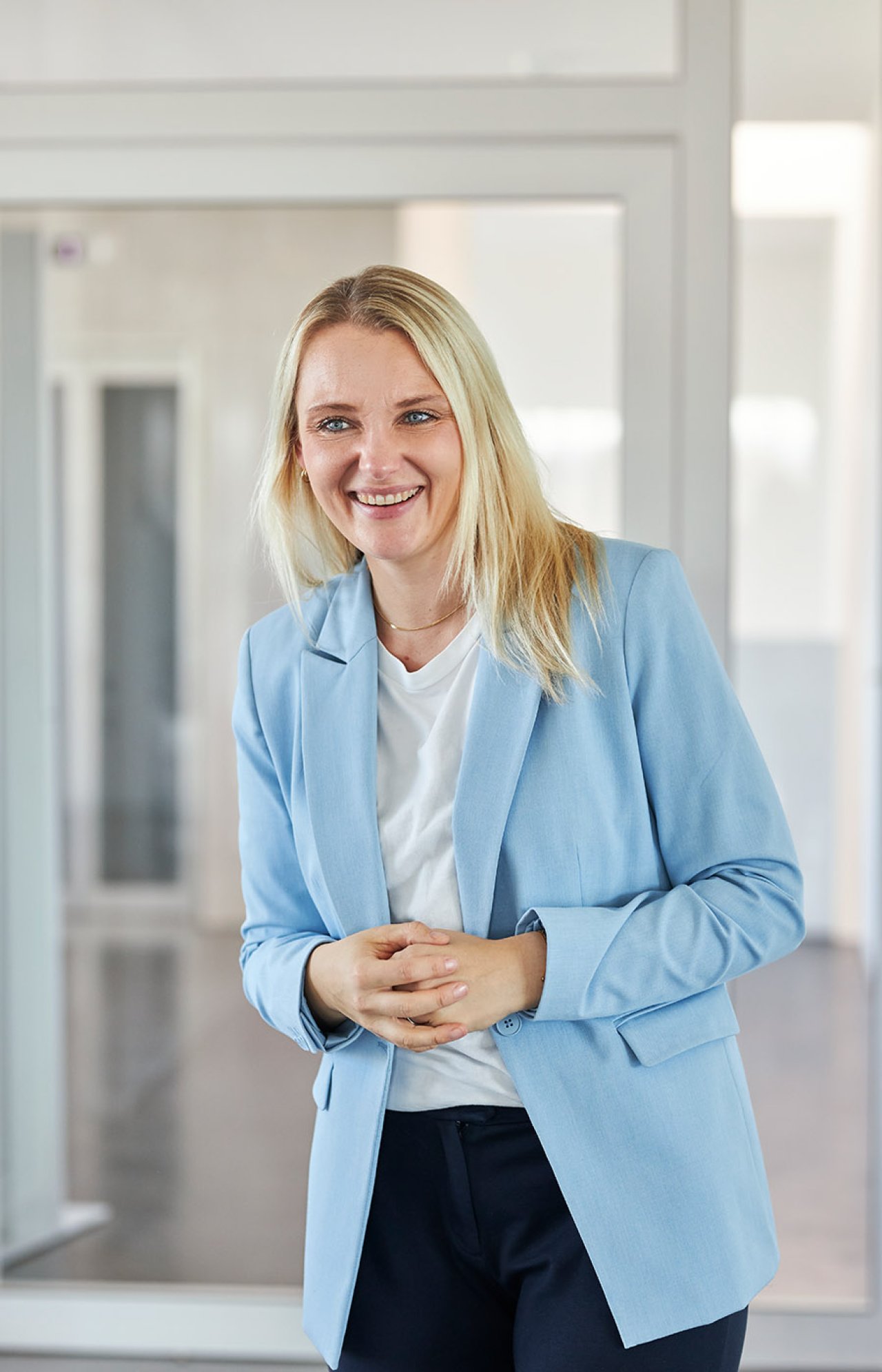 Eine blonde Frau in hellblauem Jacket steht vor einem Büro.